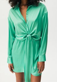 Abito camicia in satin verde con maniche lunghe, pattina bottonata, vita legata e vestibilità ampia. Texture liscia con una finitura lucida e orlo curvo.