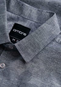 Gros plan sur le col d'une chemise grise à motif floral plié, avec une étiquette de marque noire « ombre » et une étiquette de taille M visibles.