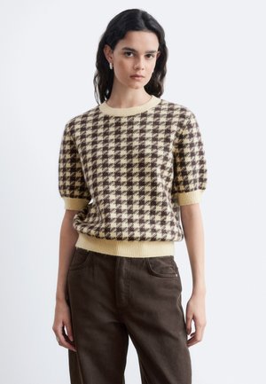 Jeune femme aux cheveux foncés portant un pull à manches courtes à carreaux beige et marron et un pantalon marron foncé sur un fond uni.
