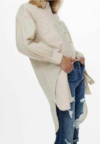 Helle beigefarbene Langarmbluse mit Knopfverschluss und Brusttaschen, kombiniert mit zerrissenen blauen Jeans. Die Bluse hat einen abgerundeten Saum.