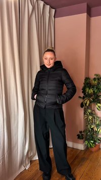 Νεαρή γυναίκα με μαύρο μπουφάν κοντά και φαρδιά μαύρη παντελόνα στέκεται μέσα σε ξύλινο πάτωμα δίπλα σε μπεζ κουρτίνα και γλάστρα με φυτό.