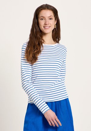 Junge Frau mit langen braunen Haaren, die goldene Creolen, ein weißes langärmliges Hemd mit blauen horizontalen Streifen und einen blauen Rock trägt.