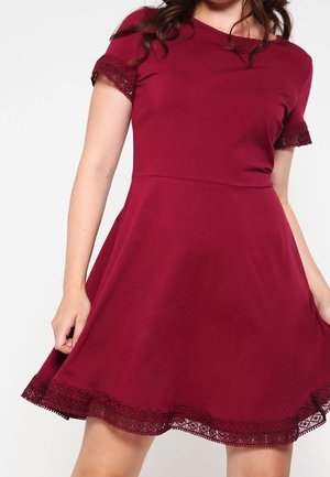 Kobieta ubrana w bordową sukienkę z krótkim rękawem, sięgającą kolan, z koronkowym wykończeniem na rękawach i dole, trzymająca boki spódnicy.