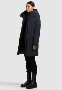 Abrigo acolchado azul marino con cuello alto, cierres de cremallera y botones, bolsillos laterales y diseño de longitud media. El modelo lleva leggings negros y botas.