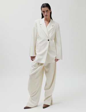 Kvinne med tilbaketrukket hår, iført en oversized kremfarget dress og brune spisse sko, stående mot en ensfarget hvit bakgrunn.