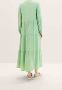 Vestido largo con volantes en tela de color verde claro. Presenta mangas largas y un escote redondeado. Combinado con zapatos beige sin hebillas.