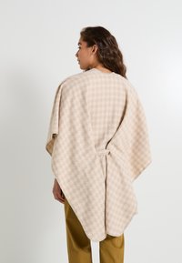 Poncho beige à motif avec une finition texturée, comportant une ceinture à la taille et des manches larges. Le design a une apparence douce et chaleureuse.