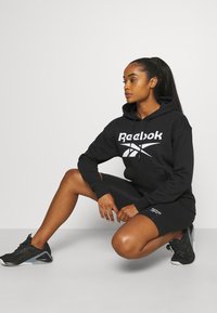 Czarna bluza z kapturem Reebok z białym logo, w połączeniu z czarnymi szortami. Model nosi czarne buty sportowe. Prosty design, materiał mieszanki bawełny.