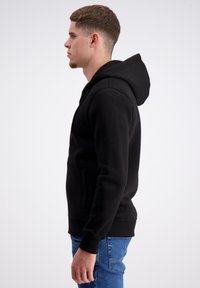 Felpa nera con tasca a marsupio, realizzata in tessuto morbido. Presenta un cappuccio con cordino e polsini e orlo a costine. Indossata con jeans blu.