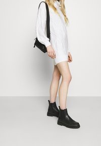 Černé kožené kotníkové boty s elastickými bočními panely a texturovanou podrážkou, spárované s bílým pleteným rolákovým šaty, které mají diamantový vzor.