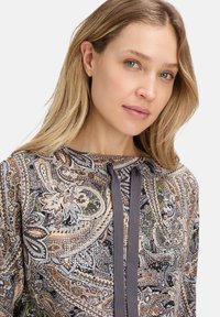 Kobieta z blond włosami w paisley'owym topie z cekinami i ciemnymi sznurkami na jasnym tle.