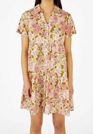 Femme portant une robe florale à manches courtes boutonnée avec des fleurs roses et vertes sur un fond clair.