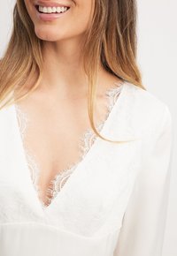 Žena v bílém topu s krajkovým výstřihem ve tvaru V, s jemným úsměvem a dlouhými světle hnědými vlasy spadajícími přes ramena.