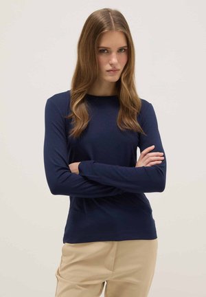 Jonge vrouw met lang golvend haar, gekleed in een marineblauw shirt met lange mouwen en beige broek, staand met gekruiste armen tegen een effen achtergrond.