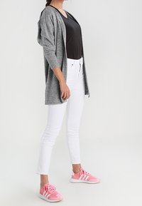 Szary dzianinowy kardigan, czarna dopasowana koszula, białe dżinsy i różowe buty sportowe z białymi akcentami. Modelka pozuje z boku, prezentując strój.