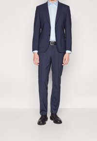 Marinblå kostym med en figursydd kavaj och raka byxor, kombinerad med en ljusblå skjorta med knappar och svart bälte, tillsammans med svarta skor.