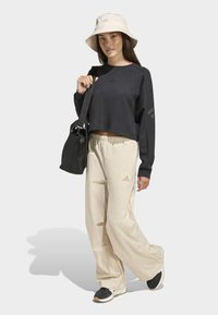 Černá krátká mikina s logem, béžové volné kalhoty, černé boty a béžový klobouk typu bucket hat. Model drží černou kabelku přes rameno.