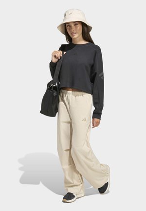 Schwarzer, kurz geschnittener Sweatshirt mit Logo, beige locker sitzende Hosen, schwarze Schuhe und beige Bucket-Hut. Das Modell hält eine schwarze Umhängetasche.