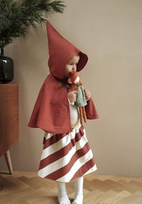 Capa de algodón roja con capucha puntiaguda y detalle de estrella, combinada con una falda de rayas en rojo y blanco. El niño sostiene una muñeca de tela.