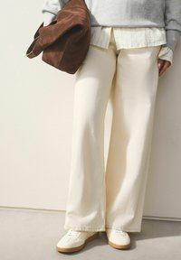 Bredde bukser i cremet farve i blødt stof med synlige syningsdetaljer. Sammen med hvide sneakers og en brun ruskindsjakke.