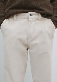 Pantalon en velours côtelé beige avec une texture côtelée, présentant des poches avant, une fermeture à bouton et un détail discret à la taille.