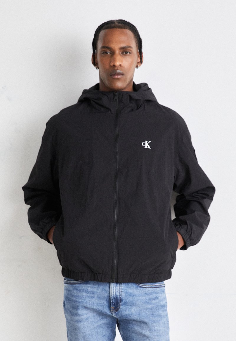 Giacca nera con cappuccio realizzata in tessuto leggero, con zip frontale, polsini elasticizzati e un piccolo logo sul petto. Indossata con jeans blu.