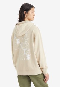 Sudadera beige con el texto "LEVIS" en blanco repetido en diversas texturas en la parte trasera. Cuenta con una capucha ajustable y un corte holgado, combinada con pantalones verdes.