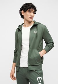 Groene hoodie met ritssluiting, een voorzak, lichtgewicht stof en een wit logo op de linkerborst, gecombineerd met een wit t-shirt met ronde hals.