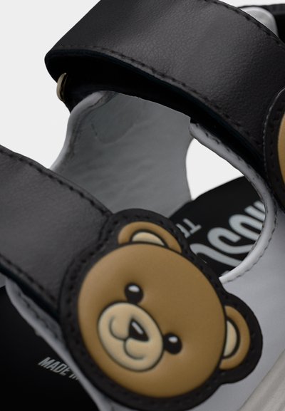 Παιδικό σανδά λι σε μαύρο και άσπρο, με χαμογελαστό πρόσωπο καφέ αρκουδάκι στο λουρί και εμφανείς ραφές στις άκρες.
