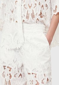 Valkoinen pitsinen asu, jossa on väljä yläosa ja elastisella vyötäröllä varustetut housut. Monimutkaisia kukkakuosisia ja läpinäkyviä materiaaleja, joissa on aaltoilevat reunat.