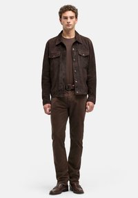 Młody mężczyzna stoi prosto, ubrany w ciemnobrązową zamszową kurtkę, dopasowane brązowe spodnie, pasek i brązowe skórzane buty na białym tle.