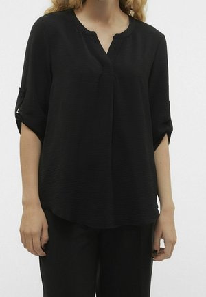 Vrouw draagt een losse zwarte blouse met opgerolde mouwen en een zwarte broek, staand tegen een effen lichte achtergrond.