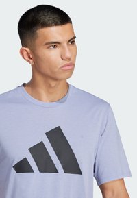 Svetlofialové tričko vyrobené z hladkej látky, s veľkým čiernym logom Adidas a tromi diagonálnymi pruhmi, s okrúhlym výstrihom a krátkymi rukávmi.