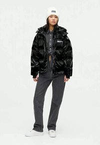 Casaco puffer preto com textura brilhante e capuz, combinado com uma camisa cinza de botões e calças cinzas. Um gorro branco e sapatilhas completam o visual.