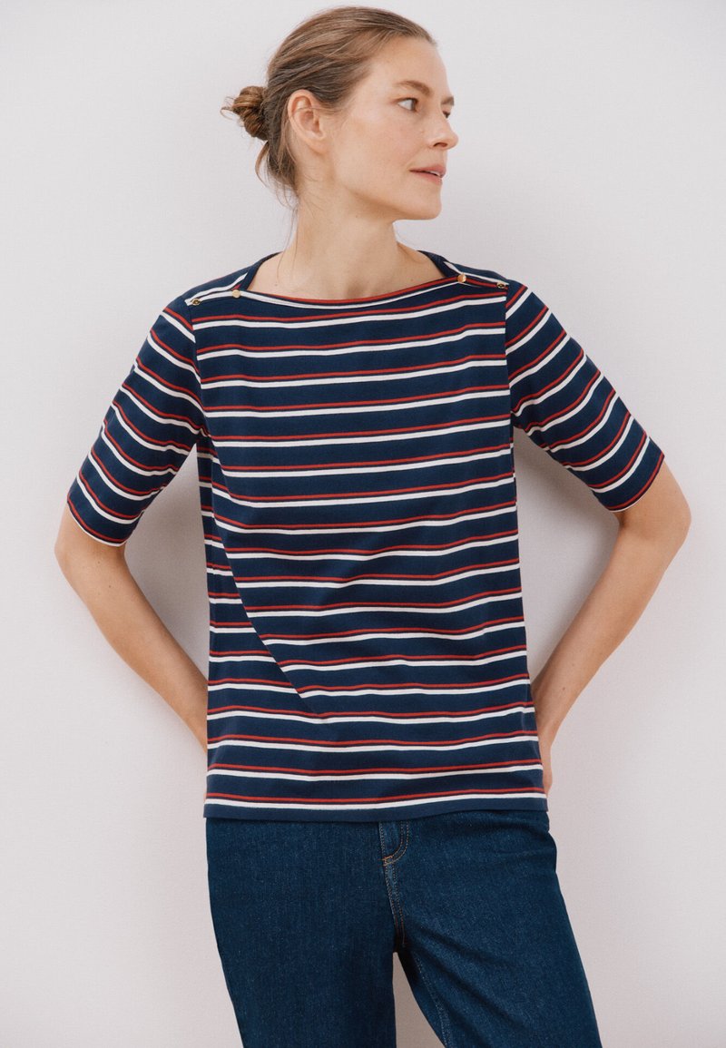 Marine stribet t-shirt med korte ærmer, røde og hvide striber, bådformet halsudskæring og dekorative knapper på venstre skulder. Stoffet er blødt.