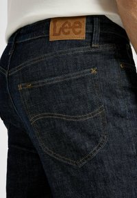 Jeans de mezclilla azul oscuro con costuras amarillas, una etiqueta de cuero marrón en la cintura y un diseño sutil en los bolsillos.