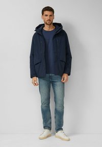 Marineblaue wasserdichte Jacke mit Kapuze, Vordertaschen und durchgehendem Reißverschluss. Wird über einem blauen Hemd getragen und mit hellen Jeans und weißen Sneakers kombiniert.