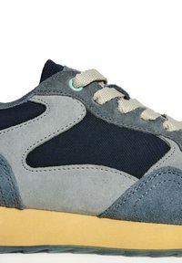 Gros plan sur une basket en daim gris et noir avec des lacets beige et une semelle en caoutchouc marron clair sur fond blanc.