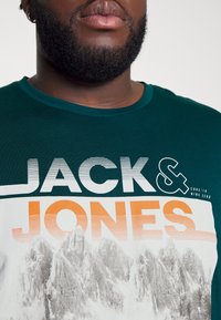 Green cotton t-shirt featuring large white and orange text, "JACK & JONES," with a graphic of mountains below. Classic crew neck design.