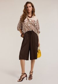 Blusa marrone con motivo, vestibilità ampia. Pantaloni corti marroni scuri con vita elasticizzata e tasca laterale. Tacchi scuri con cinturini, mentre tiene una borsa gialla.