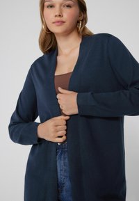 Cárdigan azul marino hecho de tejido de punto suave, con un diseño de frente abierto, mangas largas y puños acanalados, combinado con una blusa marrón.