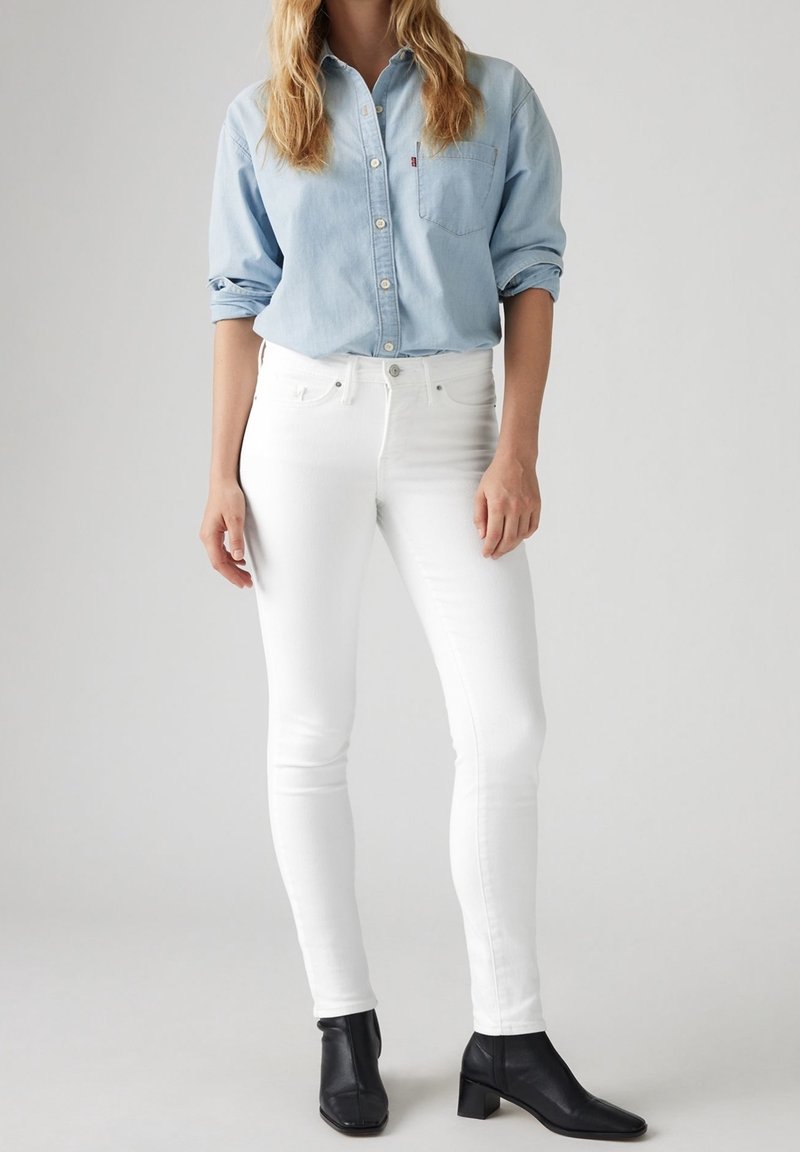 Camisa de mezclilla de color azul claro con botones y mangas enrolladas, combinada con jeans skinny blancos y botas de tacón negras hasta el tobillo.