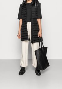 Czarna puchowa kamizelka z kapturem, wykonana z teksturowanej tkaniny, noszona na czarnym t-shircie. Do tego kremowe spodnie o szerokich nogawkach i czarne botki. W zestawie czarna torba typu tote.
