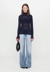 Top din tricot cu guler înalt de culoare bleumarin, purtat cu blugi evazați de un albastru deschis. Modelul ține o geantă din piele neagră, stând pe un fundal simplu.