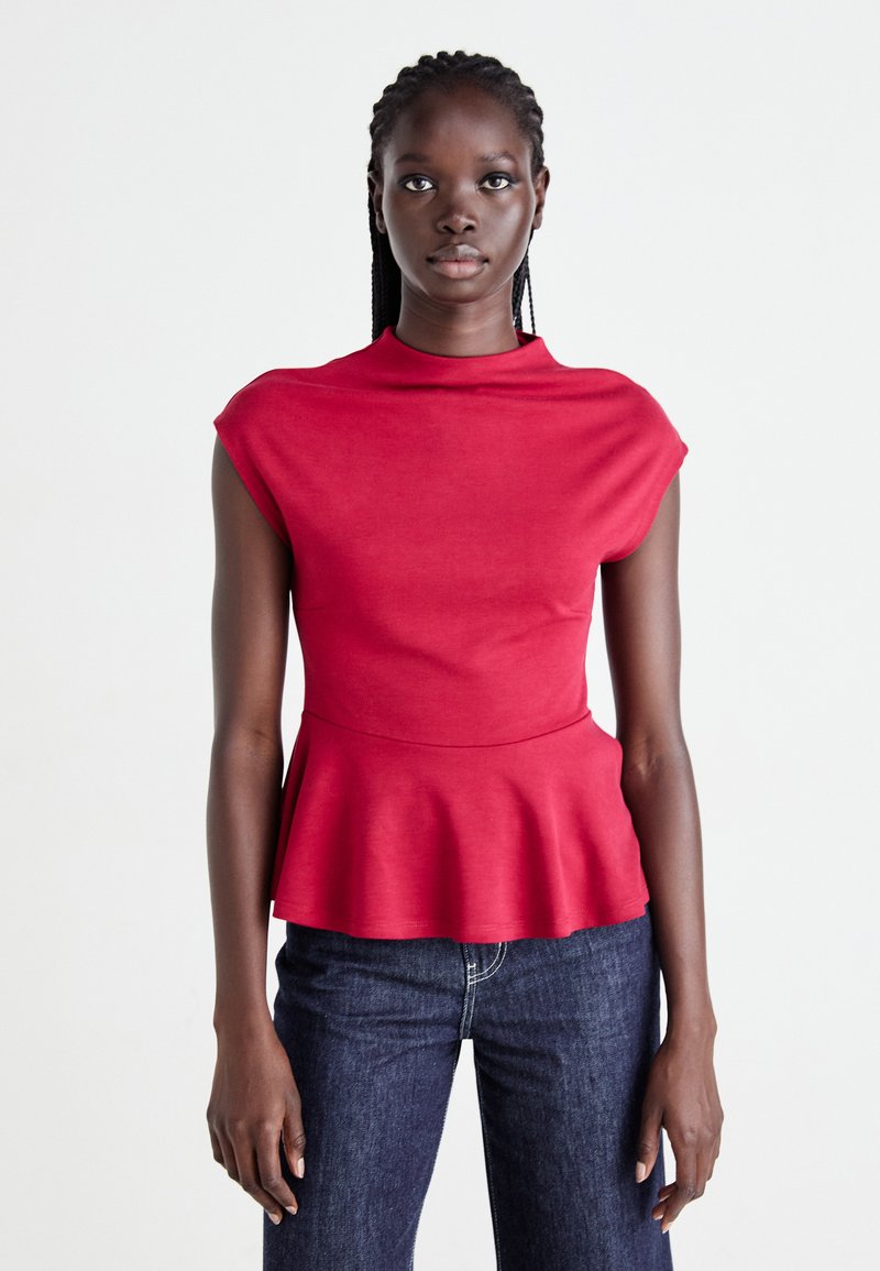 Kvinne med flettet hår som har på seg en ermeløs rød peplum-topp og mørkeblå jeans, som står foran en ensfarget hvit bakgrunn.