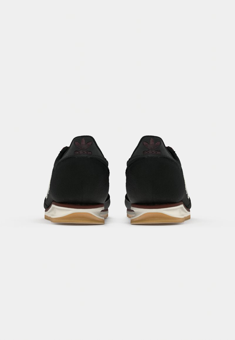 Zapatillas deportivas negras con parte superior texturizada, suela de goma y acentos marrones. Presenta el logo de adidas en los talones y bordes de suela de color claro.