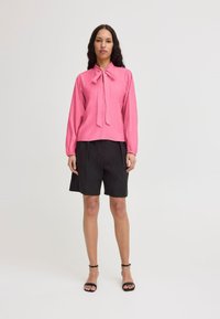 Blusa rosa a maniche lunghe con colletto a fiocco, abbinata a pantaloni corti neri. Texture liscia, vestibilità rilassata; sandali con cinturini alla caviglia.
