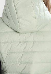 Lysegrøn quiltet jakke med hætte båret af en person set bagfra, skulderlangt hår synligt.