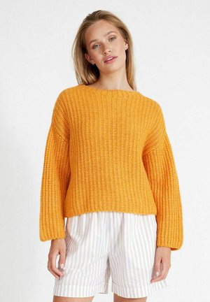 Fleckiger Strickpullover in leuchtendem Orange, mit weitem Halsausschnitt, langen Ärmeln und gerippter Textur. Kombiniert mit gestreiften Shorts in Weiß und Beige.
