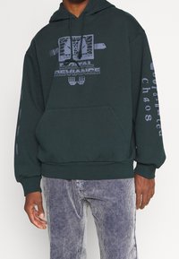 Tmavě zelená mikina s kapucí, na přední straně s grafikou a nápisem "Digital Deviance", dlouhými rukávy s potištěným textem a klokaní kapsou.